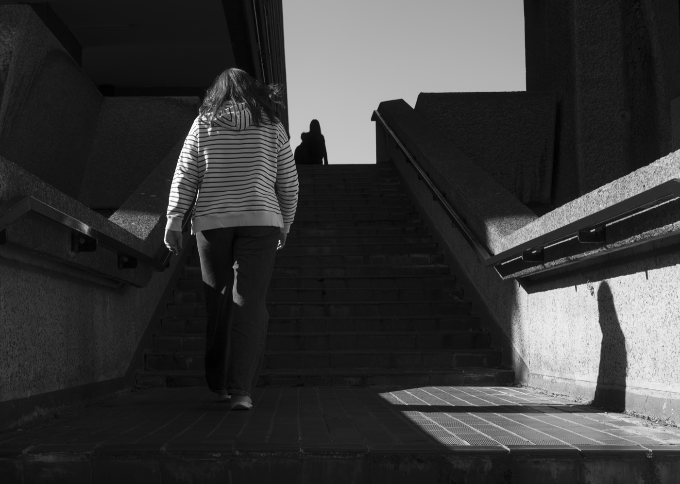 A figure climbing a Brutalist concrete staircase that opens into bright light at the top