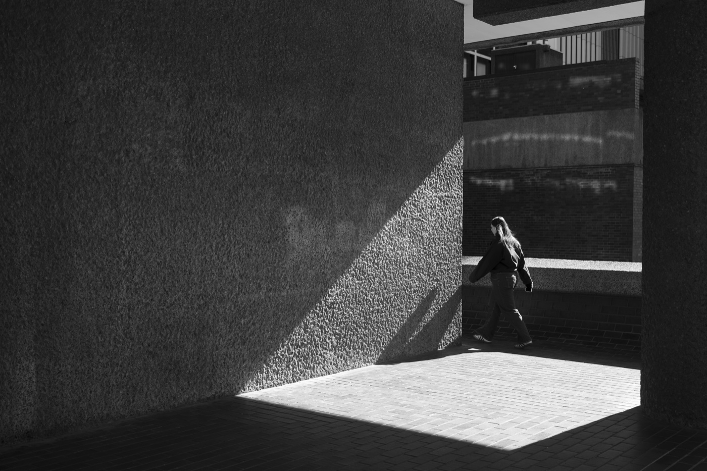 A figure walking through a wedge of light cast by a concrete wall on the Barbican podium