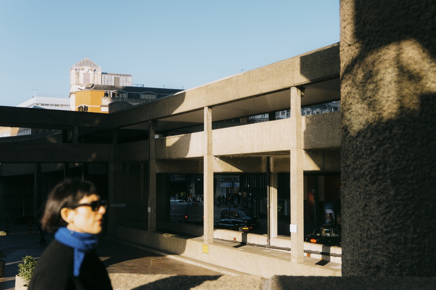 A woman in a blue scarf walking through warm afternoon light at the Barbican